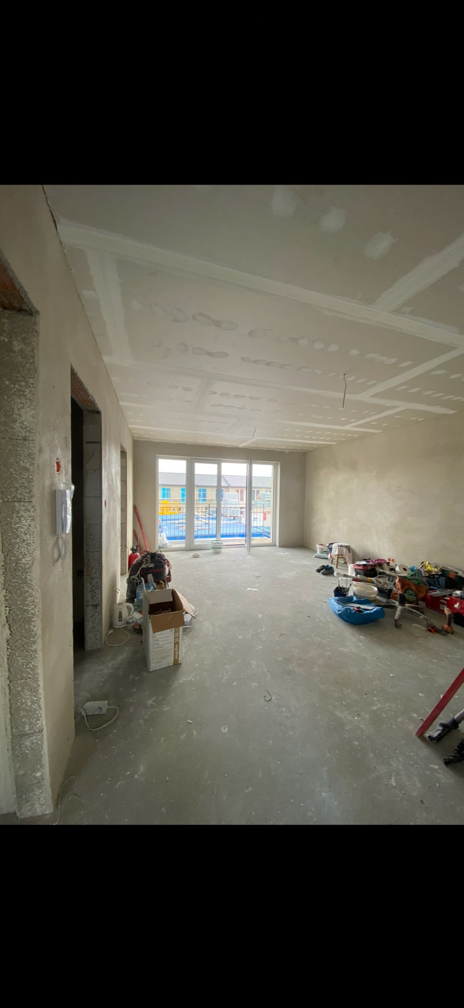 Surowy stan wnętrza mieszkania w trakcie prac wykończeniowych: ściany i sufit pokryte płytami kartonowo-gipsowymi, widoczne szpachlowanie łączeń, podłoga betonowa, okno balkonowe z widokiem na nowe...