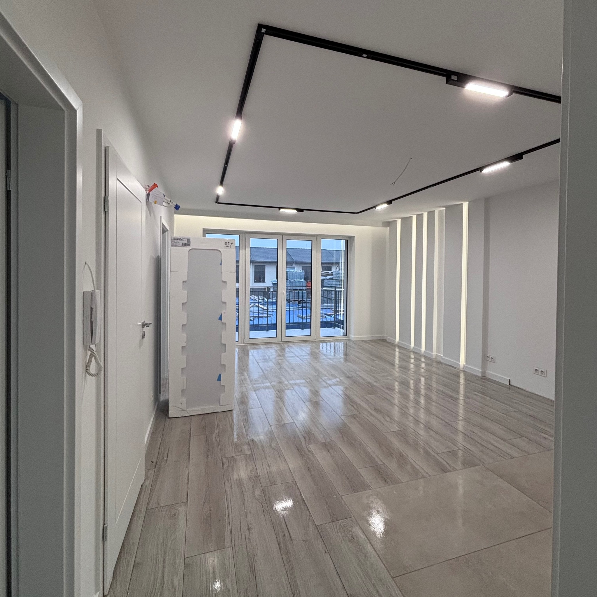 Jasne, przestronne wnętrze mieszkania z panelami podłogowymi imitującymi drewno, oświetleniem szynowym na suficie i dużymi oknami balkonowymi wychodzącymi na osiedle.