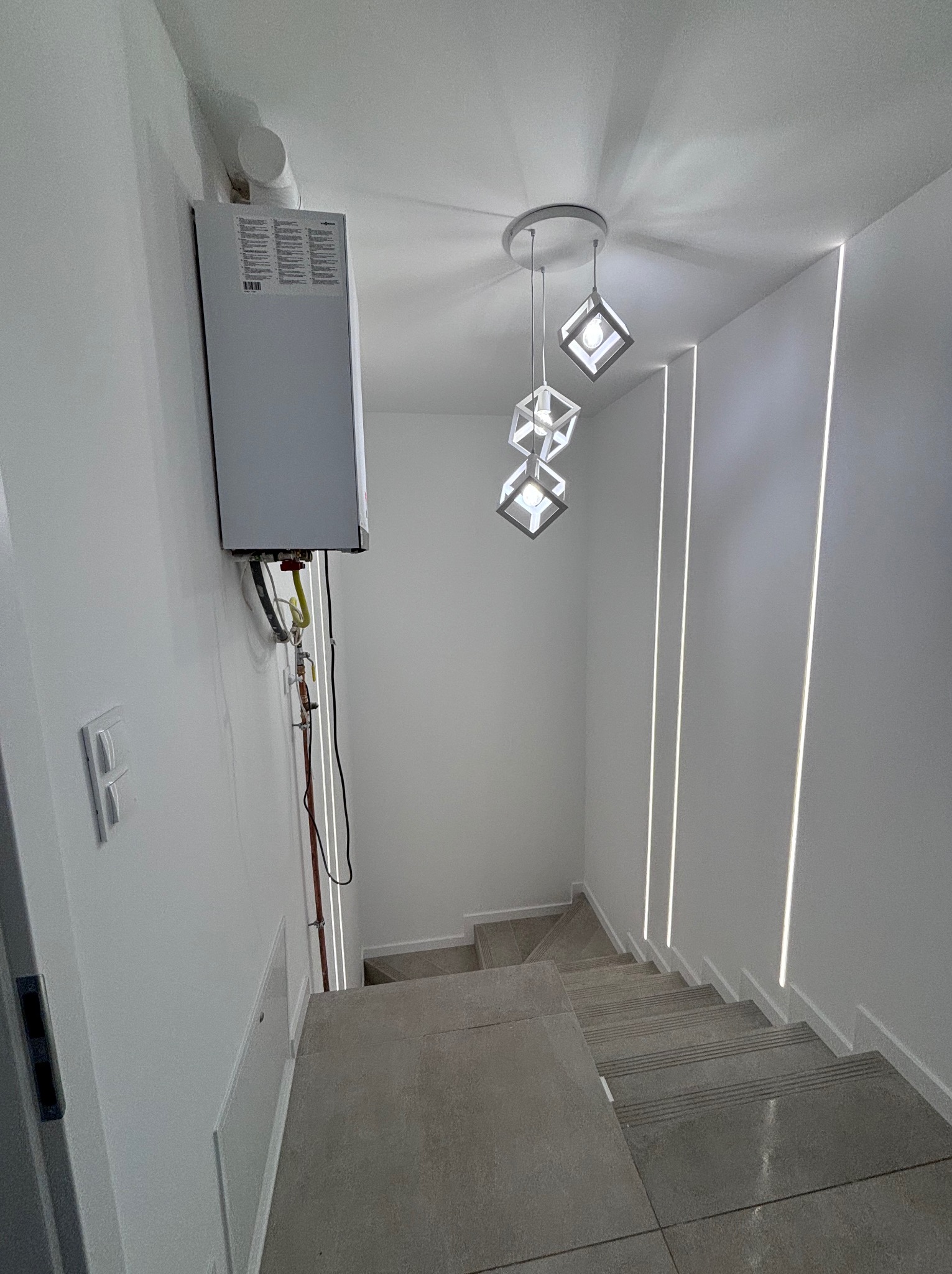 Klatka schodowa z szarymi płytkami, oświetlona nowoczesną lampą wiszącą z geometrycznymi kloszami i pionowymi pasami światła w ścianie, z widocznym piecem gazowym na ścianie.