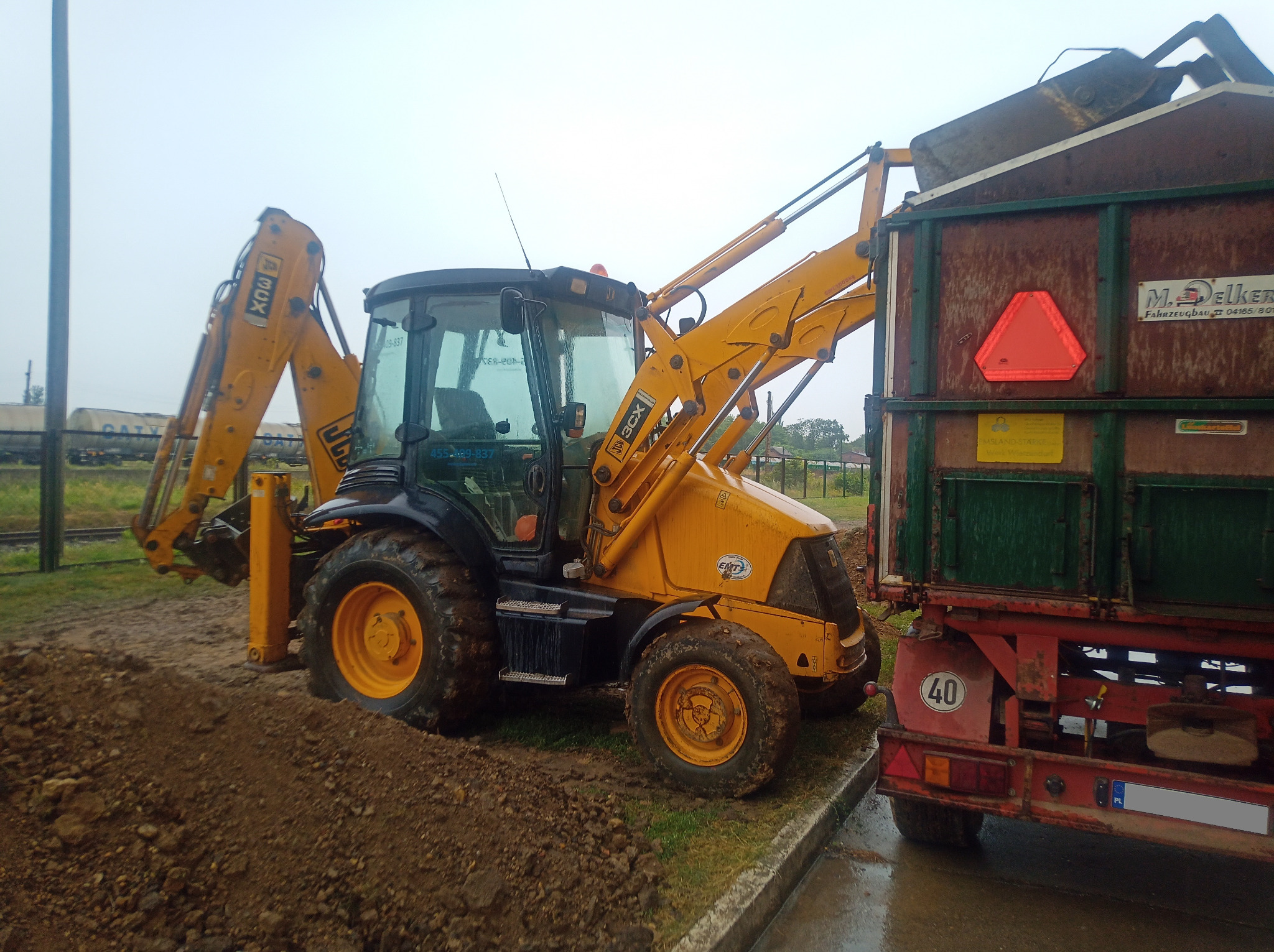 Żółta koparko-ładowarka JCB 3CX z łyżką nad wywrotką, obok pryzma ziemi, widoczne tory kolejowe w tle.