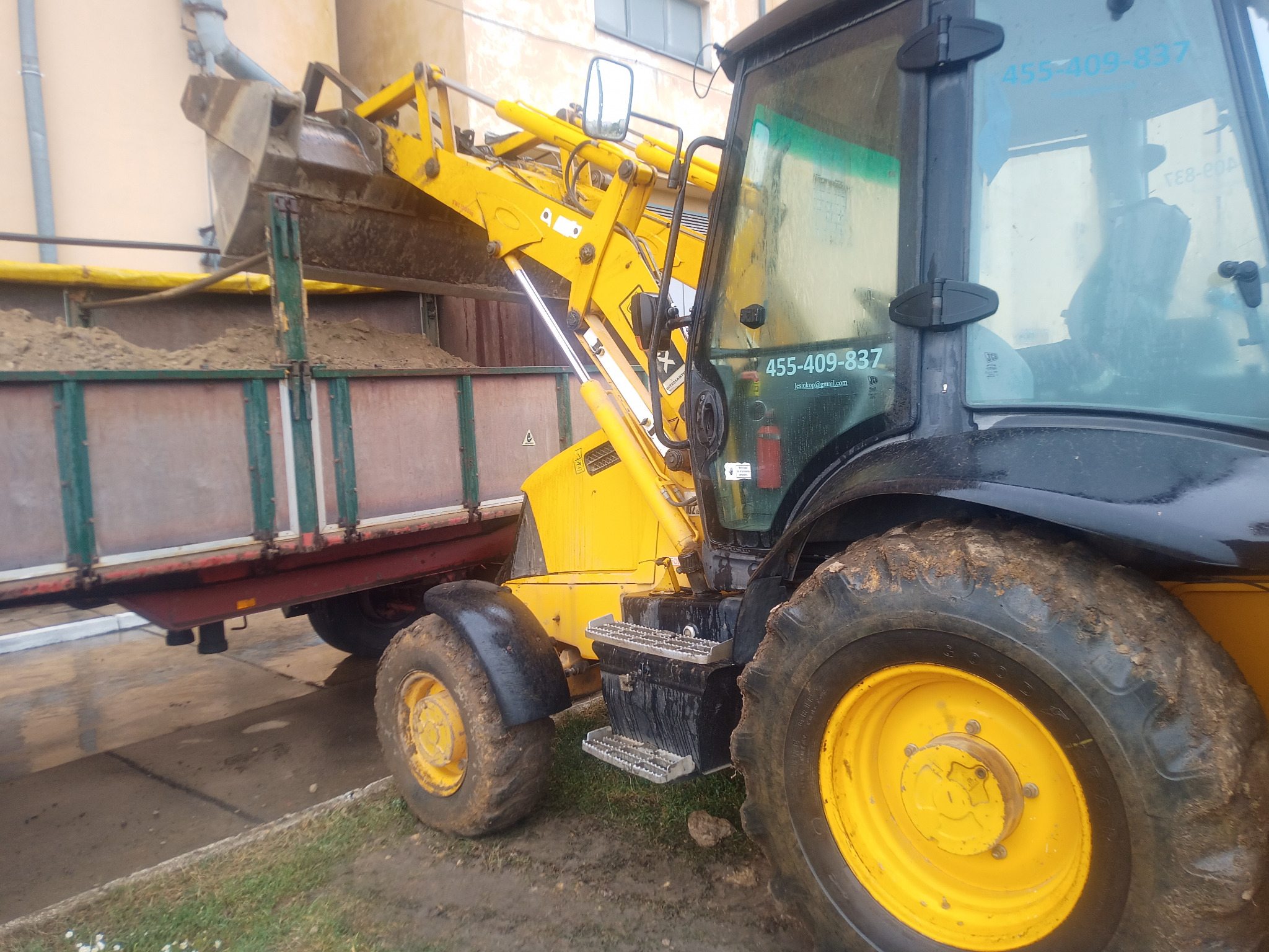 Żółto-czarna koparko-ładowarka z łyżką załadowaną ziemią, wysypuje urobek do przyczepy, widoczne błotniste koła maszyny i numer kontaktowy na szybie kabiny.