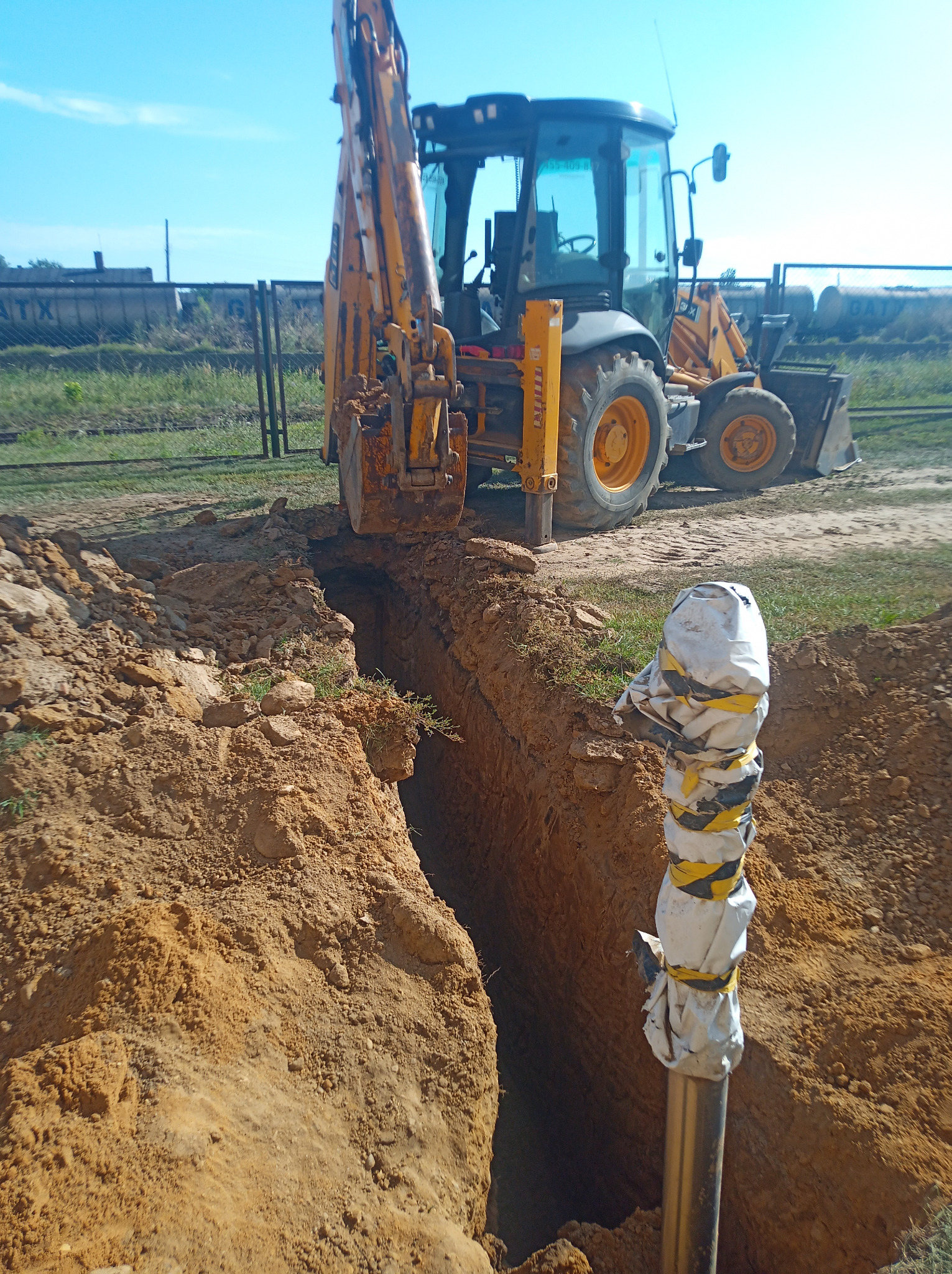 Żółta koparko-ładowarka w trakcie wykonywania wąskiego wykopu w ziemi, z widoczną rurą zabezpieczoną folią i taśmą ostrzegawczą, sugerującą prace instalacyjne.