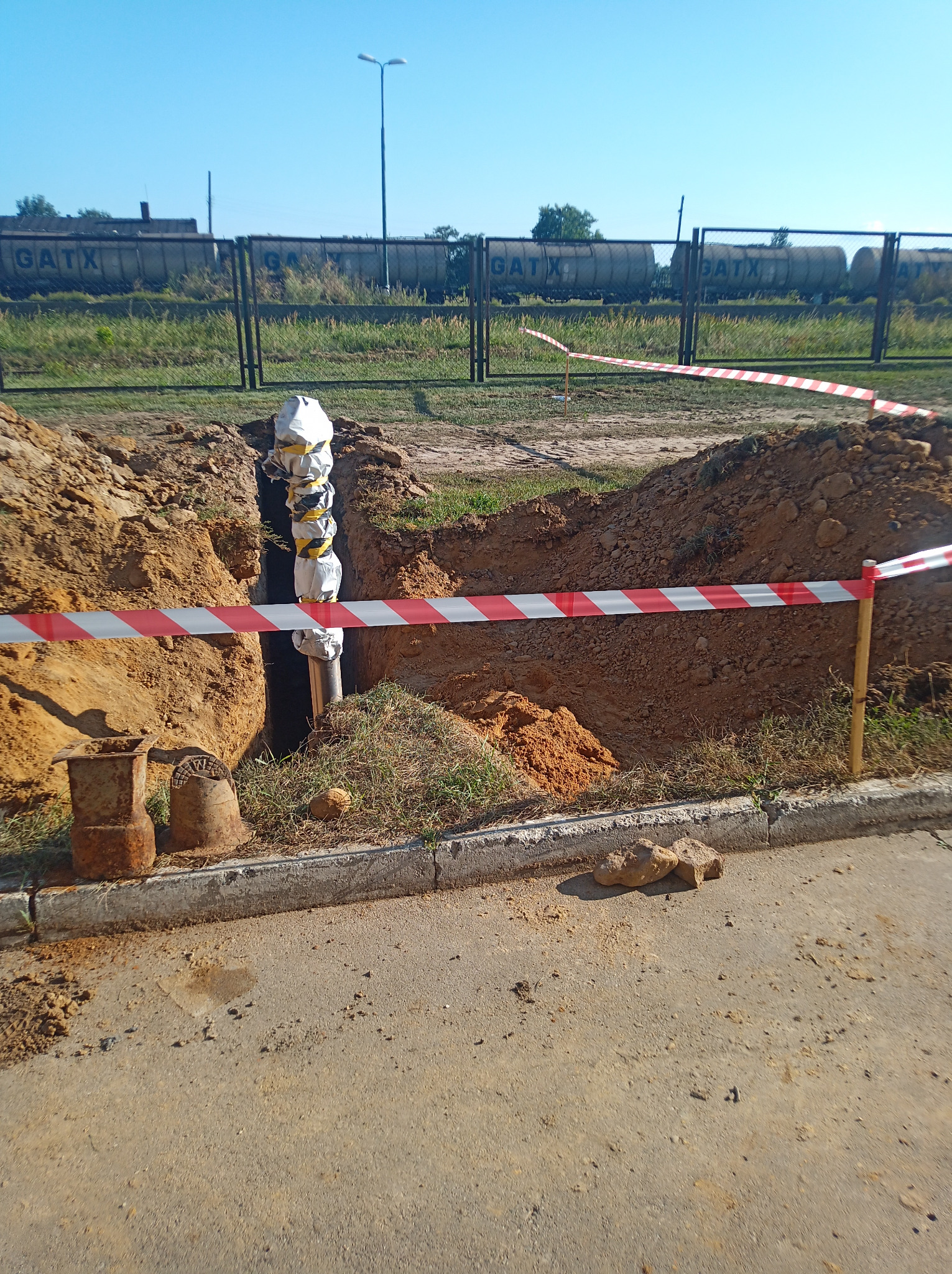 Wykop w ziemi z rurą owiniętą izolacją, zabezpieczony taśmą ostrzegawczą, widoczne metalowe elementy i tory kolejowe w tle.