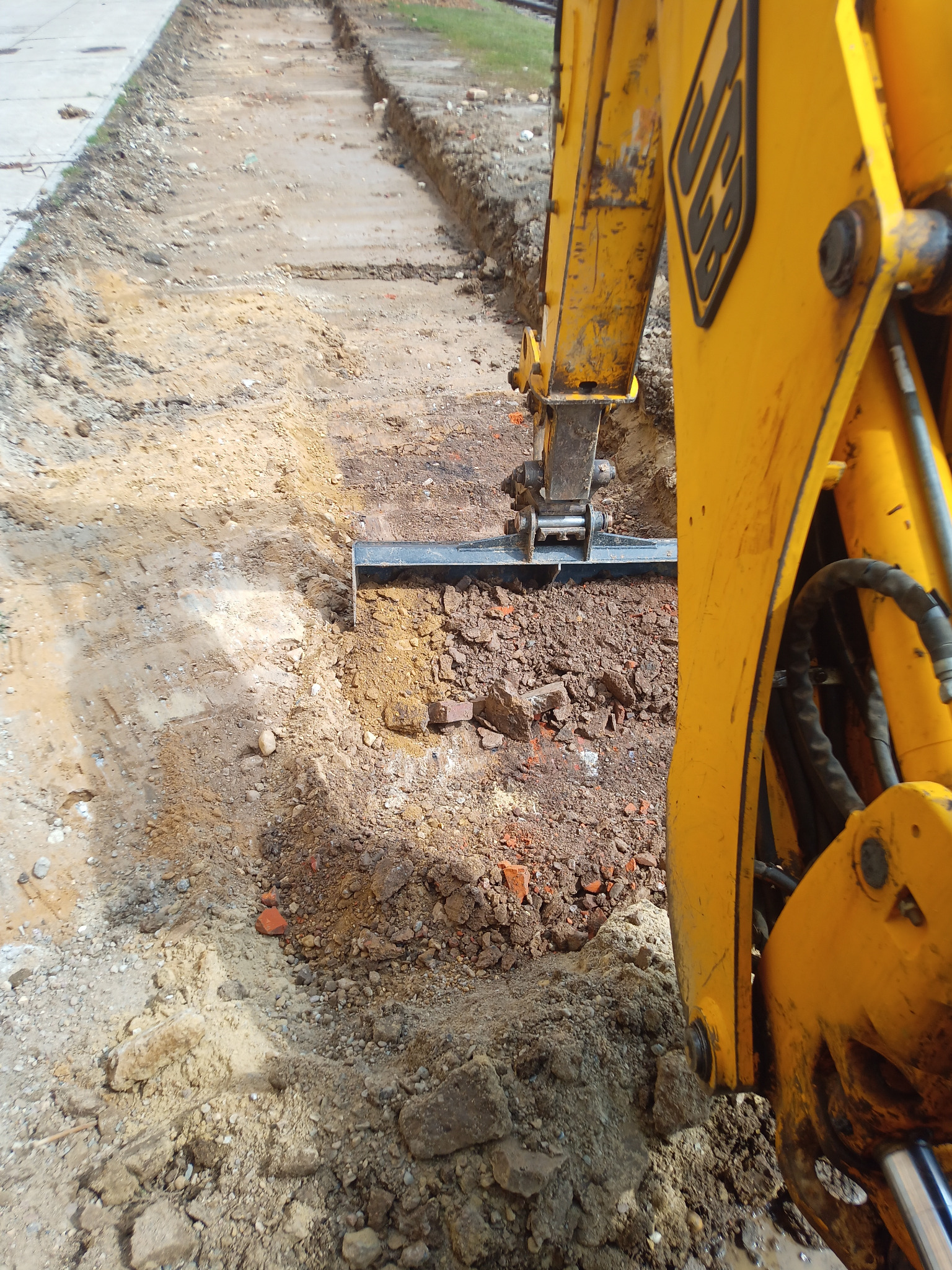 Żółta koparka JCB wyrównuje wąski wykop w ziemi z gruzem ceglanym, widok z góry na roboczą część maszyny i krawędź wykopu.