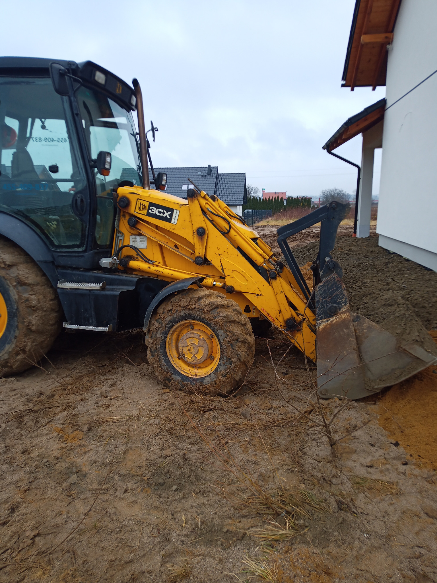 Żółta koparko-ładowarka JCB 3CX w trakcie prac ziemnych przy budynku. Widoczne błoto na kołach i świeżo usypana ziemia.
