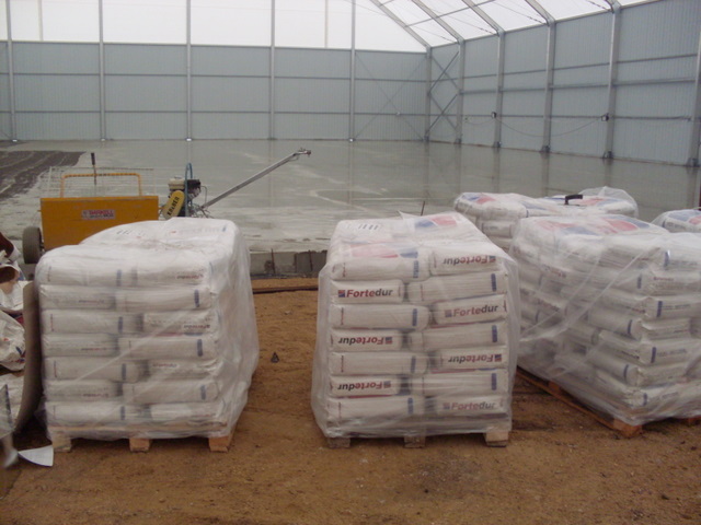 Wnętrze hali w Zagrodnie z przygotowanymi materiałami budowlanymi Fortedur na paletach, gotowymi do wykonania posadzki, w tle widoczna maszyna do wylewania.