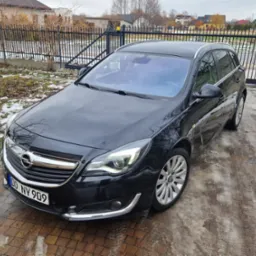 Czarna Opel Insignia kombi zaparkowana na brukowanym podjeździe z resztkami śniegu, widoczna tablica rejestracyjna 'DO NY 909', w tle metalowe ogrodzenie i domy.