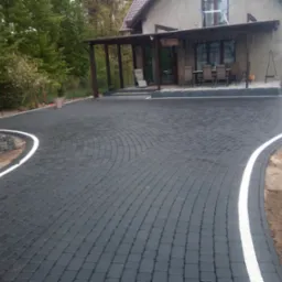 Świeżo ułożona, ciemnoszara kostka brukowa z białymi obrzeżami prowadząca do domu z zadaszonym tarasem i meblami ogrodowymi.