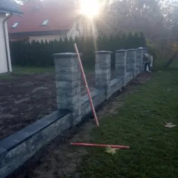 Montaż modułowego ogrodzenia z szarej kostki łupanej; widoczny pracownik w trakcie murowania i poziomica oparta o słupek; w tle dom z czerwoną dachówką i iglaki.