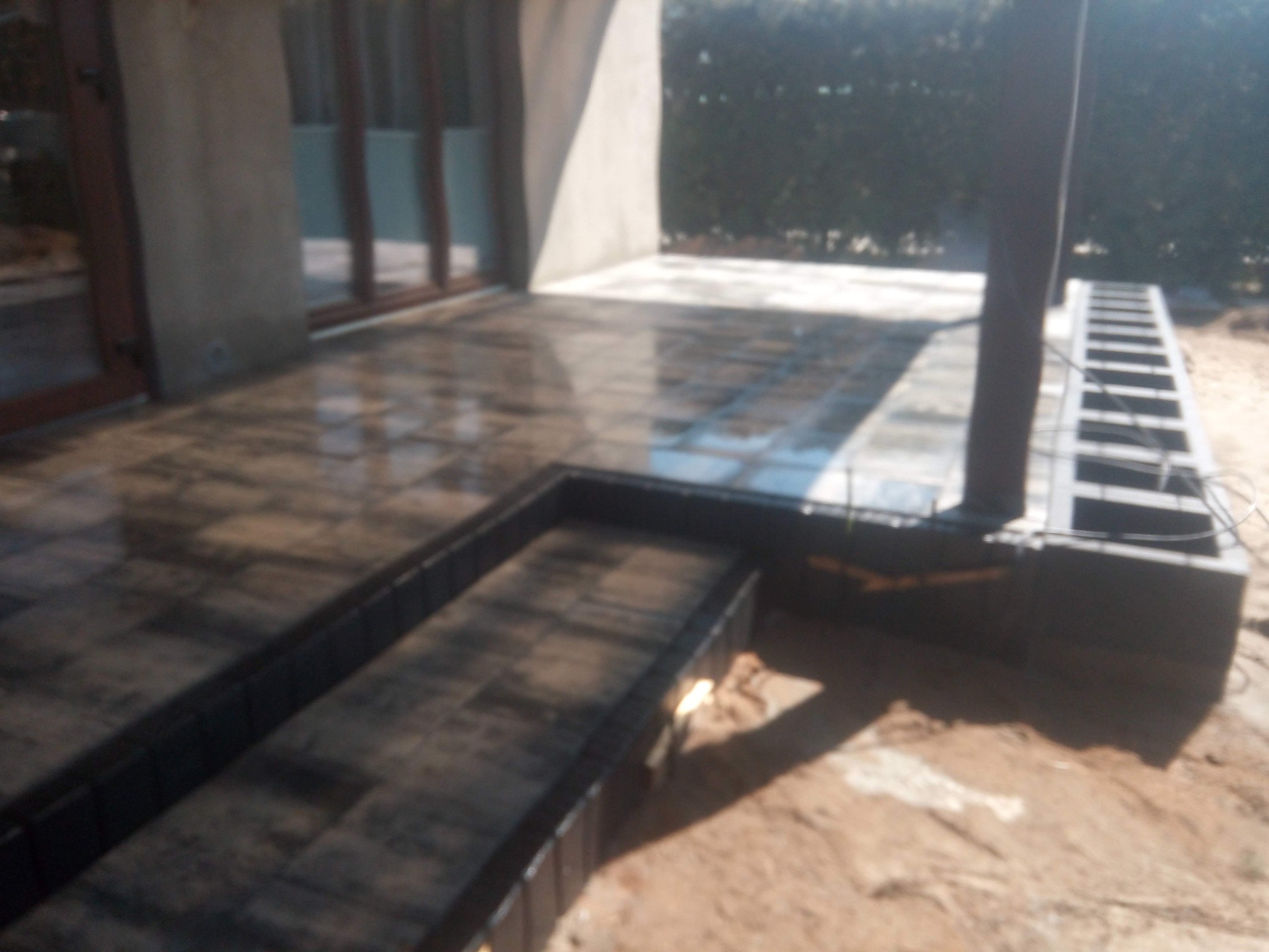 Ułożona, błyszcząca kostka brukowa na tarasie przylegającym do budynku, z widocznym obrzeżem i przygotowanym miejscem na schody, w tle zieleń.