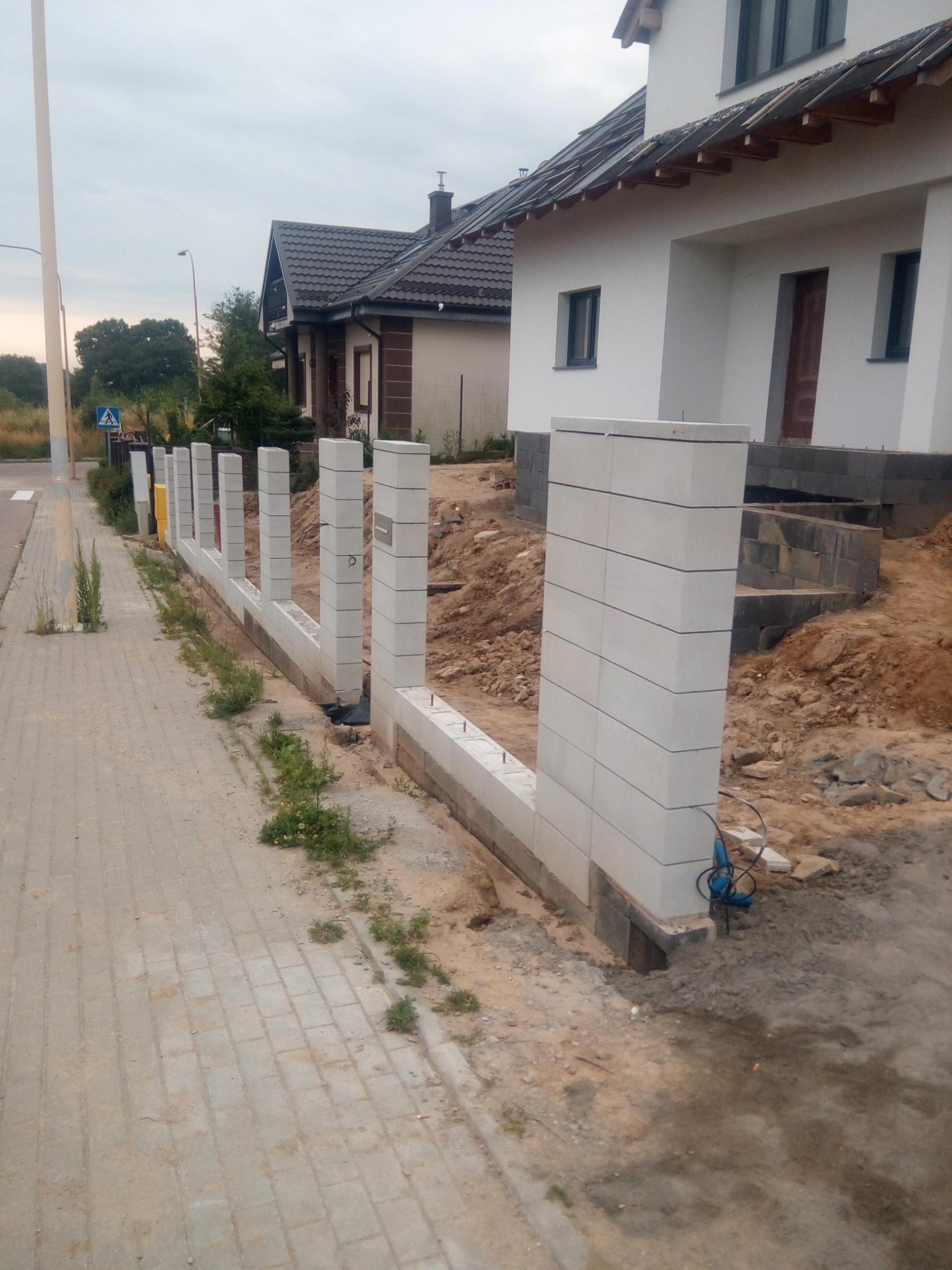 Wznoszenie ogrodzenia z bloczków betonowych przy chodniku i domu w trakcie budowy, widoczne elementy fundamentu i narzędzia.