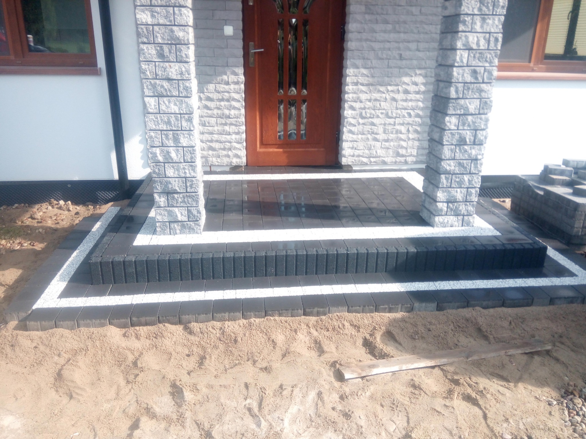 Nowo ułożony chodnik z kostki brukowej prowadzący do wejścia domu, z widocznym piaskiem i niezakończonymi pracami wykończeniowymi wokół.