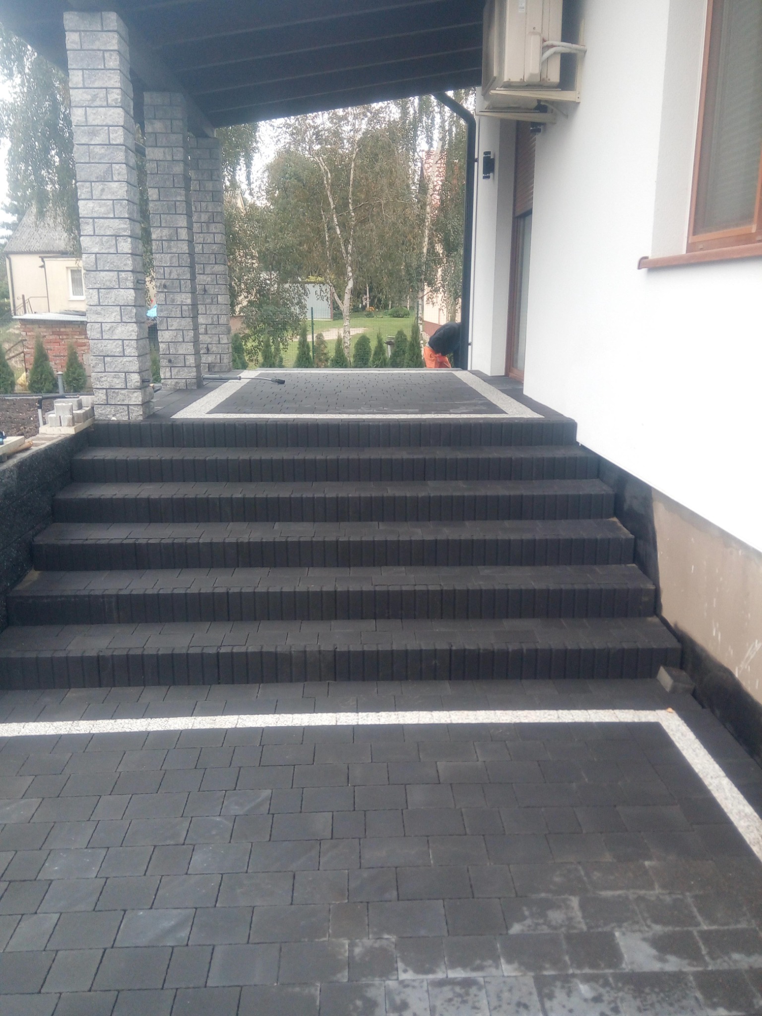 Schody wejściowe do domu wyłożone ciemną kostką brukową z białymi akcentami, widoczne kolumny z szarej cegły i fragment elewacji budynku.