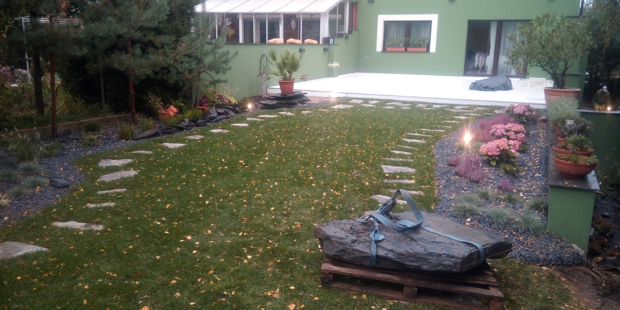 Ogród z ścieżką z kamiennych płyt na trawniku pokrytym jesiennymi liśćmi, z dekoracyjnym żwirem, kwiatami i oświetleniem, duży kamień na palecie w pierwszym planie.