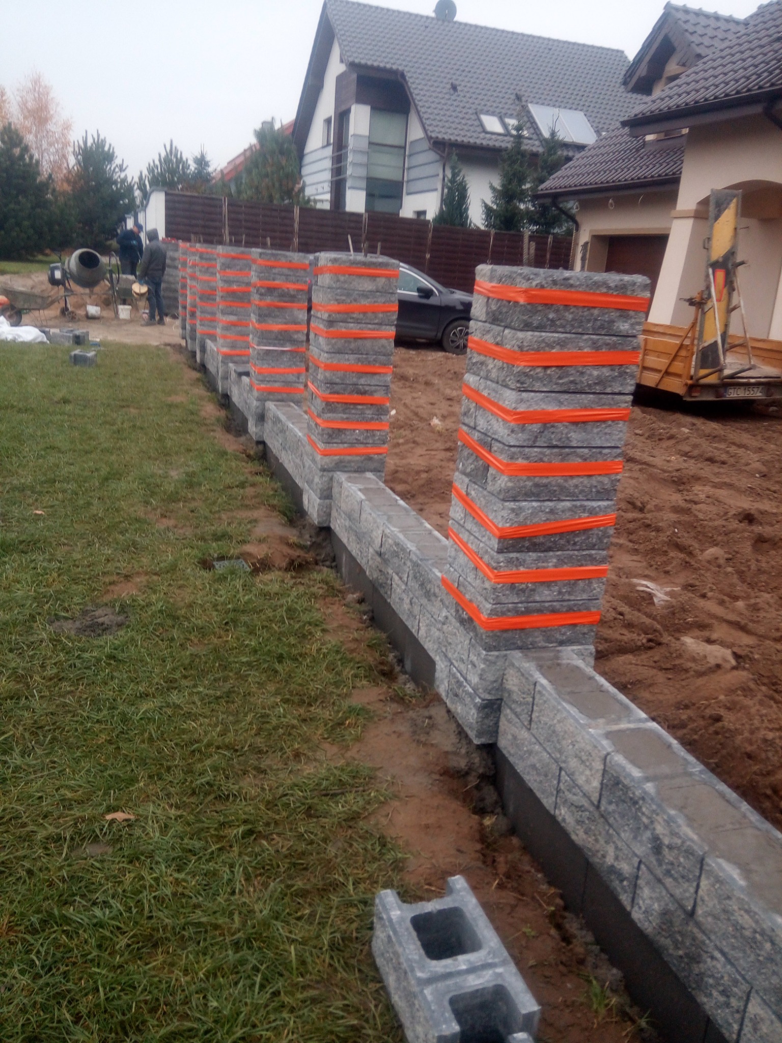 Budowa ogrodzenia z szarej kostki, słupki zabezpieczone pomarańczową taśmą, w tle dom jednorodzinny i betoniarka.