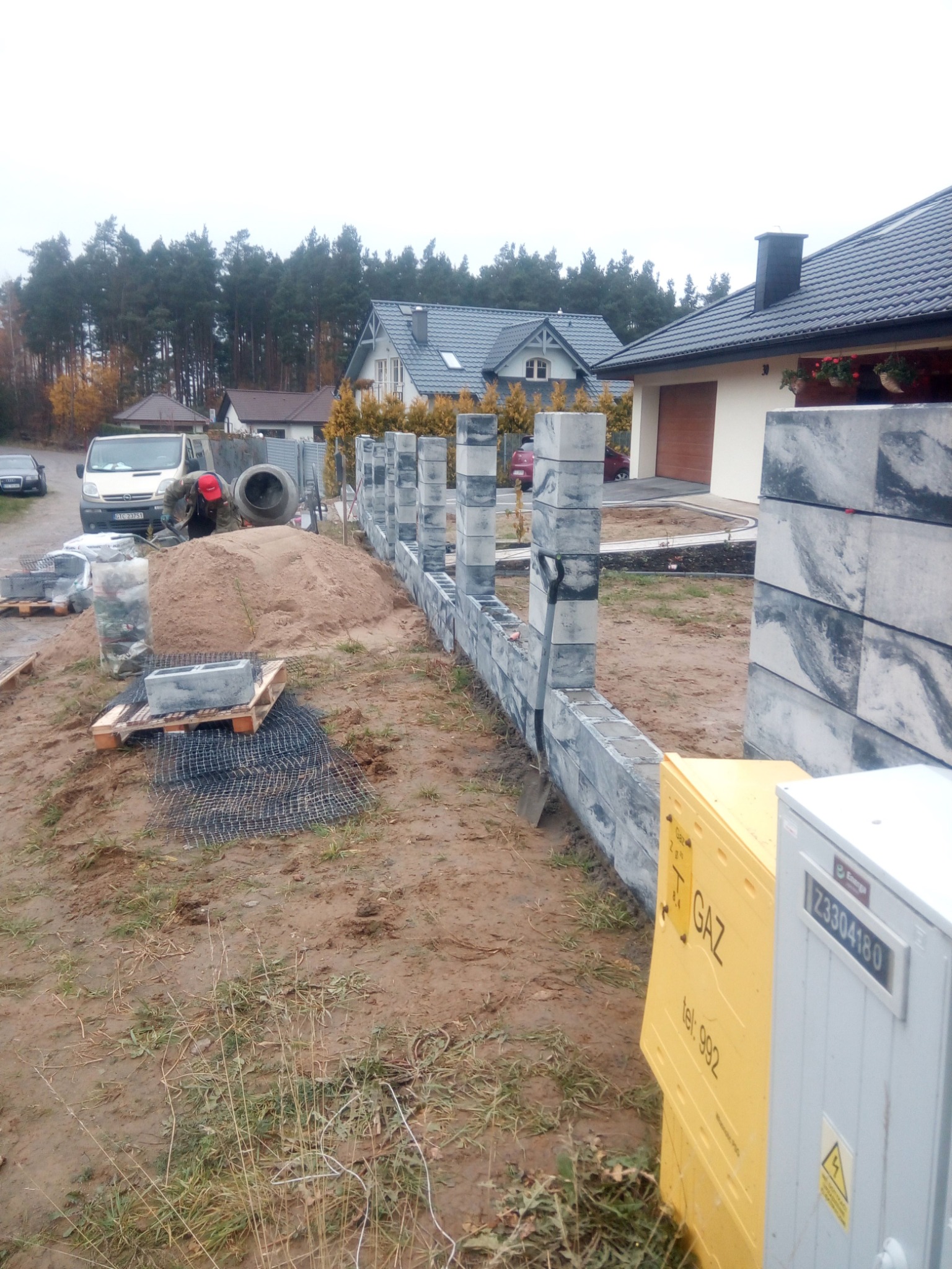 Budowa ogrodzenia z szarej kostki modułowej; widoczny fragment muru, słupki, palety z kostką, betoniarka, piasek, narzędzia i pracownik w czerwonej czapce