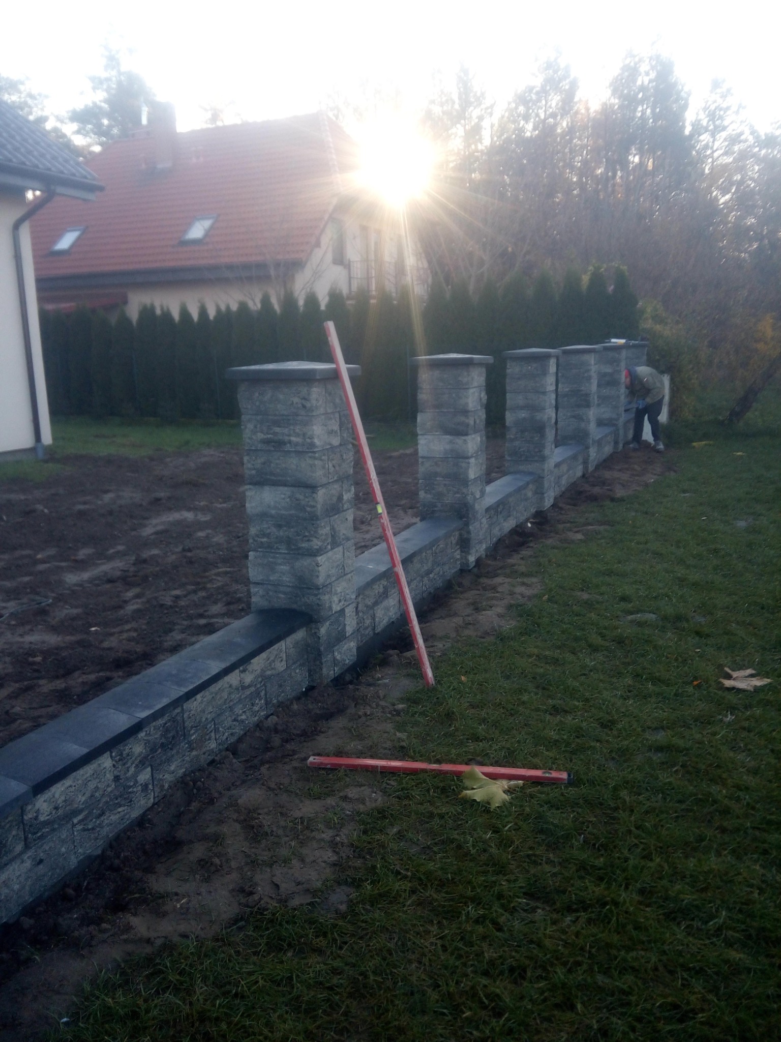 Montaż modułowego ogrodzenia z szarej kostki łupanej; widoczny pracownik w trakcie murowania i poziomica oparta o słupek; w tle dom z czerwoną dachówką i iglaki.