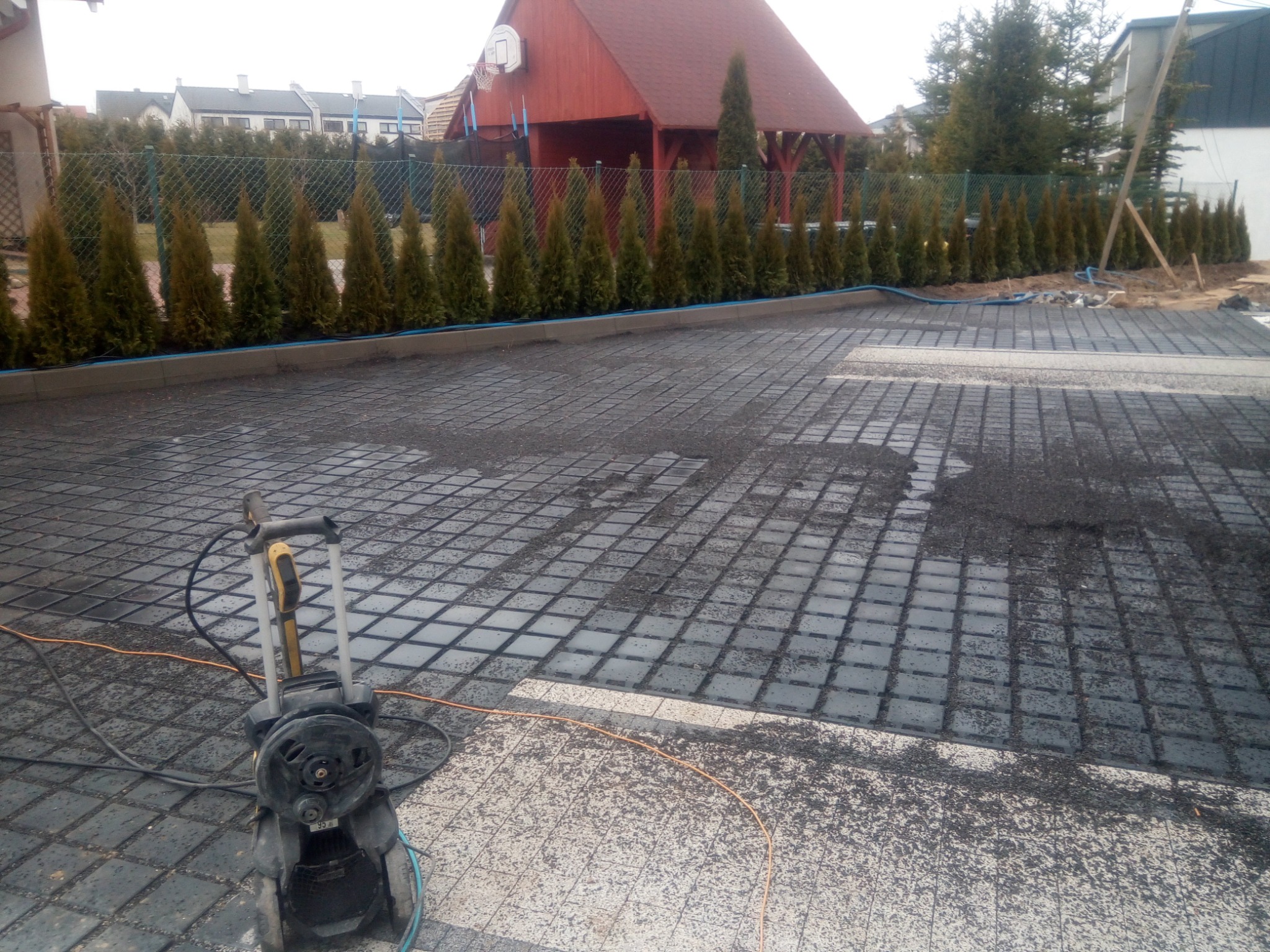 Przygotowany podkład pod kostkę brukową z widoczną siatką stabilizującą, obok myjka ciśnieniowa, w tle rząd tuj i zabudowa jednorodzinna.