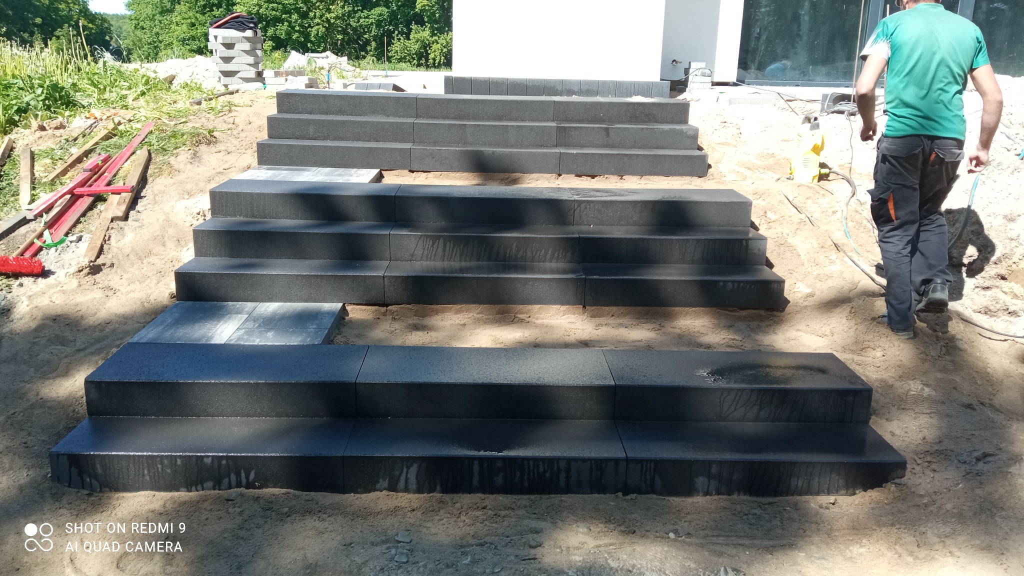 Schody zewnętrzne z ciemnej kostki brukowej w trakcie budowy, widoczny pracownik w tle, teren wokół w trakcie przygotowania.