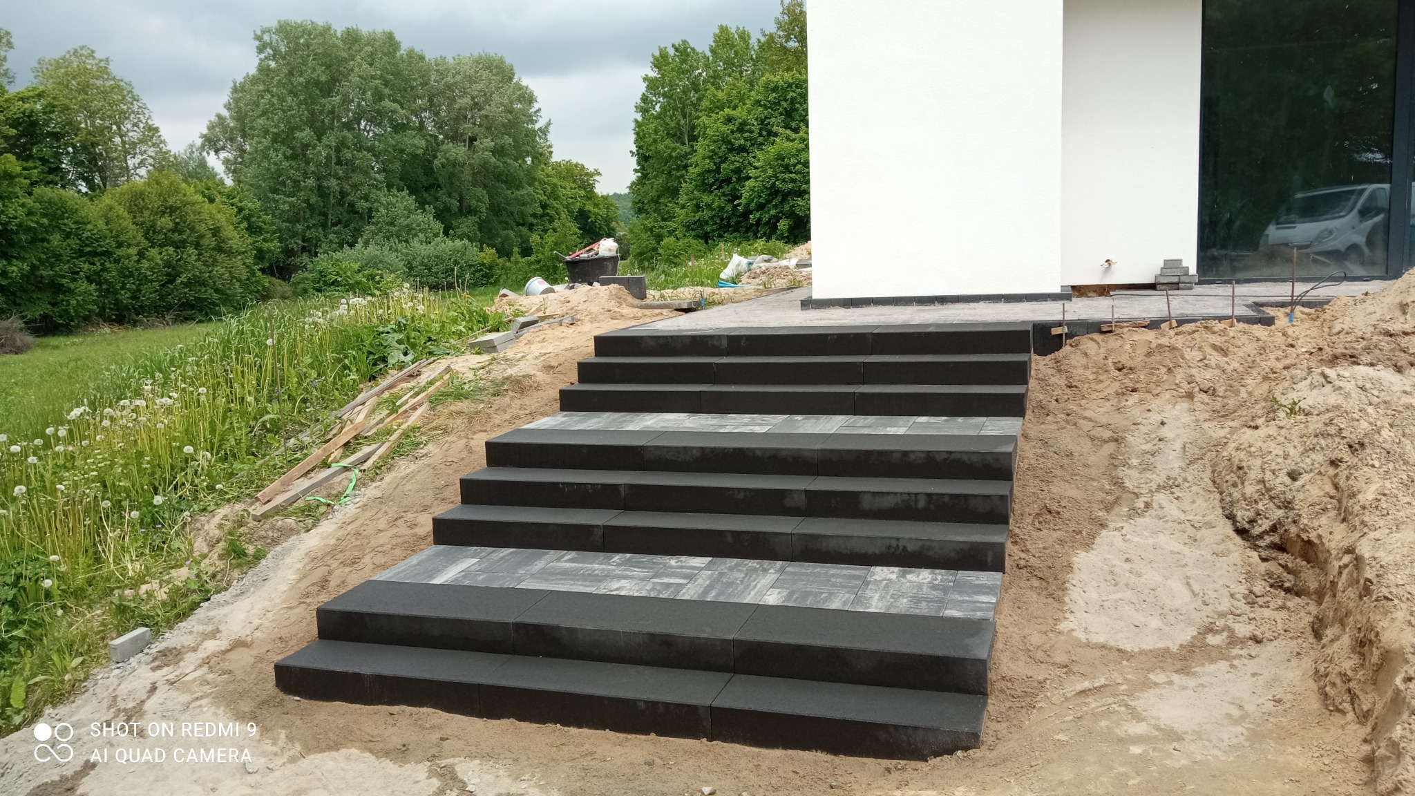 Schody zewnętrzne z szarej kostki brukowej prowadzące do nowoczesnego budynku z białymi ścianami i dużym oknem, teren wokół w trakcie prac ziemnych.