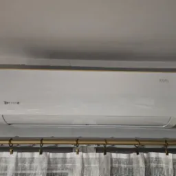 Zamontowany klimatyzator Rotenso Roni Series, widoczny z dołu, z zasłoniętą częścią dolną przez białe, lniane zasłony na złotym karniszu.