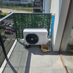 Elektro-Usługi Bartłomiej Pawlaczyk - Biała jednostka zewnętrzna klimatyzacji Bosch na balkonie, z żółtym wiaderkiem obok, widok na zewnątrz z balustradą z sztucznych liści.