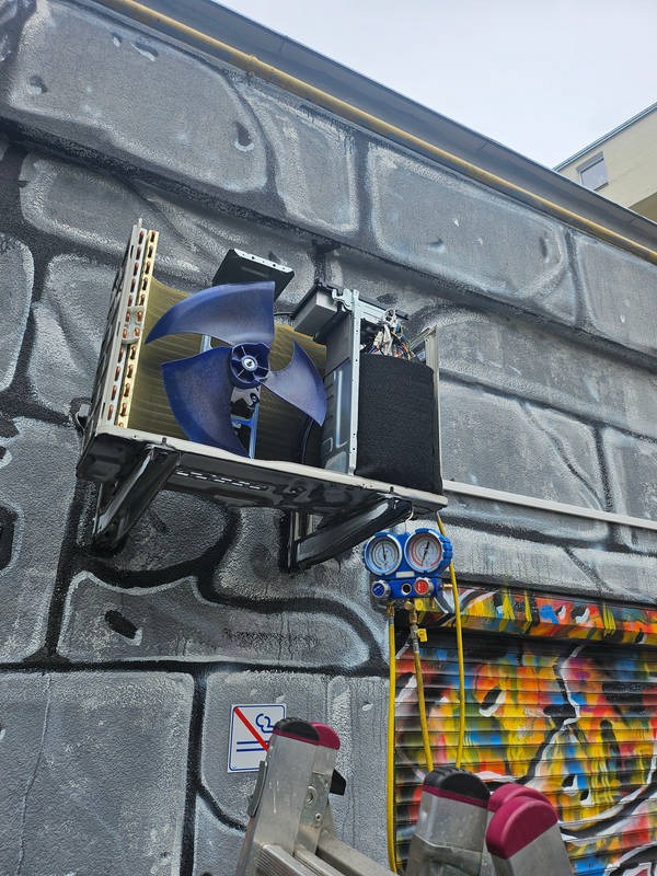Demontaż klimatyzatora zewnętrznego na ścianie pokrytej graffiti, widoczne manometry i drabina.
