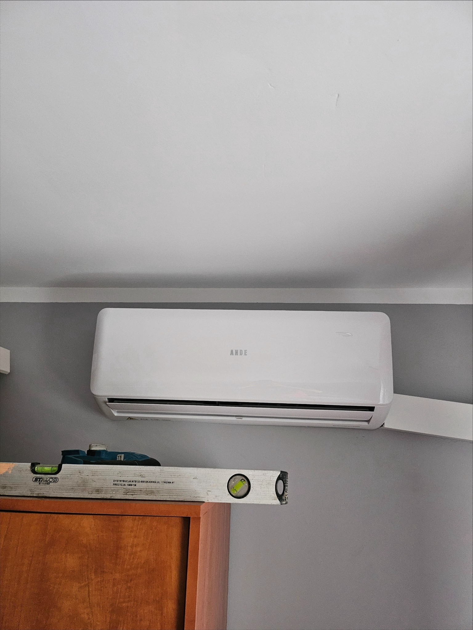 Biały klimatyzator marki ANDE zamontowany na szarej ścianie, poziomica STALCO leży na szafce pod urządzeniem, sprawdzając jego wypoziomowanie.