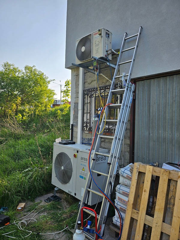 Montaż klimatyzacji typu split z jednostką zewnętrzną Toshiba na ścianie budynku, widoczna drabina oparta o ścianę, narzędzia i materiały montażowe wokół, trawa i paleta w tle.