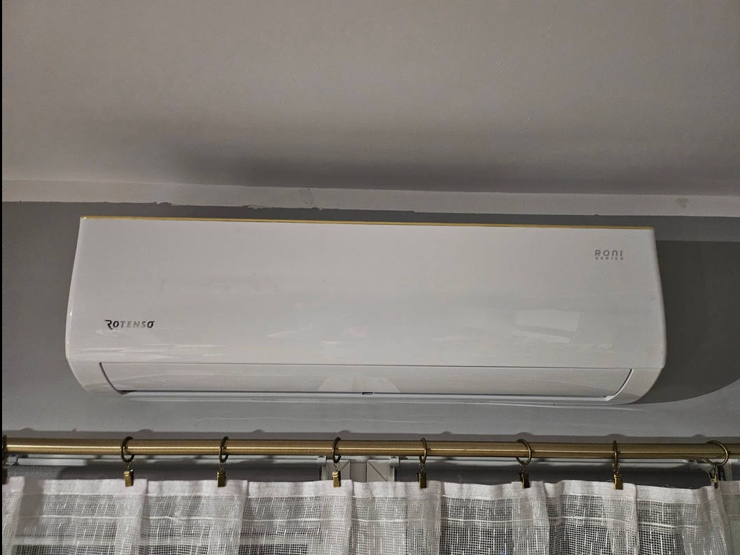 Zamontowany klimatyzator Rotenso Roni Series, widoczny z dołu, z zasłoniętą częścią dolną przez białe, lniane zasłony na złotym karniszu.