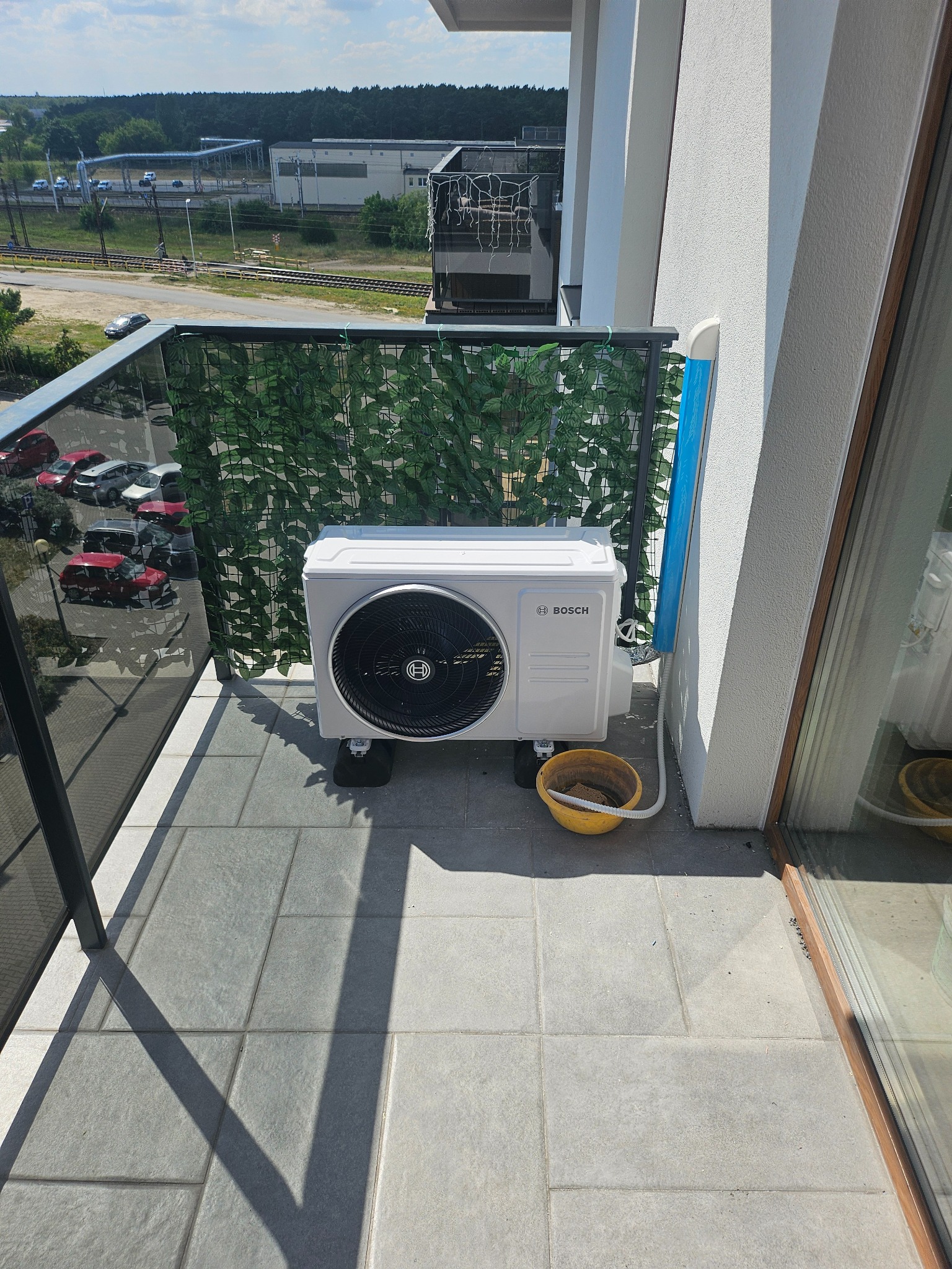Biała jednostka zewnętrzna klimatyzacji Bosch na balkonie, z żółtym wiaderkiem obok, widok na zewnątrz z balustradą z sztucznych liści.