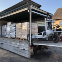 Otwarta paka samochodu dostawczego, załadowana paletami z workami i kostkami opału, obok wózek widłowy z silnikiem spalinowym.