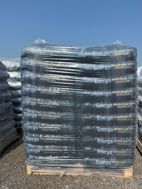 Paleta czarnych worków z ekogroszkiem marki Energo, zabezpieczonych folią, na tle błękitnego nieba.