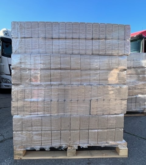 Paleta brykietu drzewnego, zabezpieczona folią stretch, widoczna na tle zaparkowanych ciężarówek w słoneczny dzień.