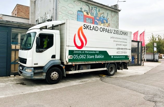 Biała ciężarówka z logo i danymi kontaktowymi składu opału 'Skład Opału Zielonki' na tle budynku z muralem.