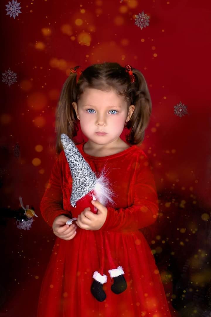 Portret dziewczynki w czerwonej sukience trzymającej ozdobnego skrzata na tle czerwonego tła z motywem śnieżynek.