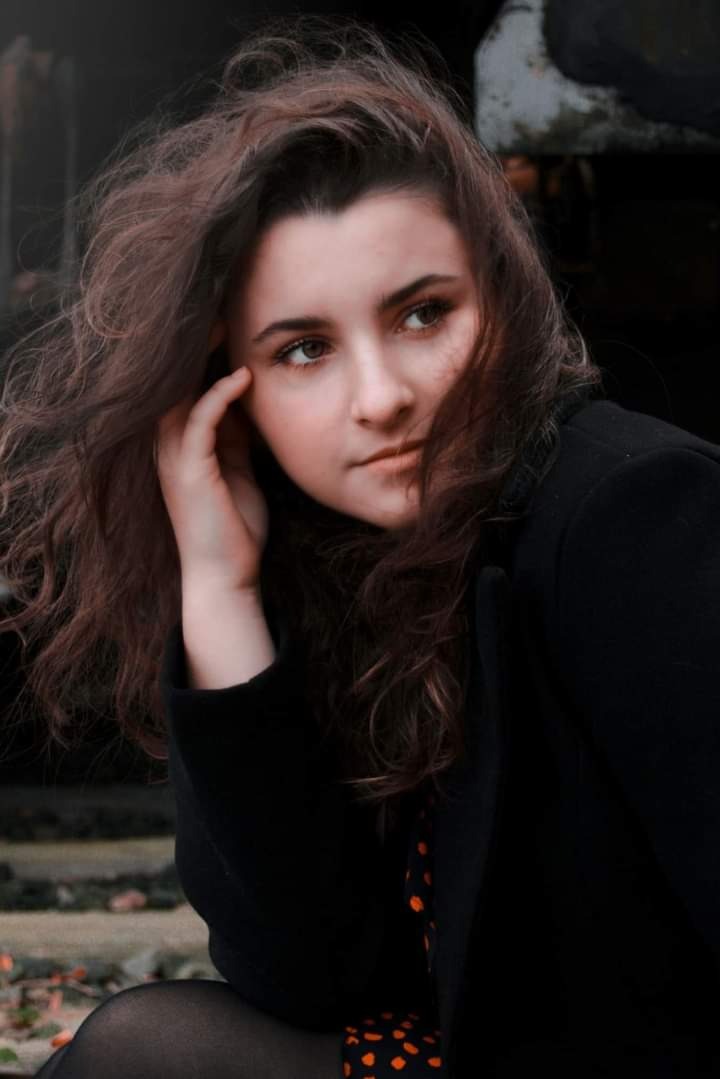 Portret młodej kobiety z rozwianymi brązowymi włosami, w czarnym płaszczu i sukience w pomarańczowe kropki, wykonany w Przemyślu na tle kamiennych schodów i jesiennych liści.