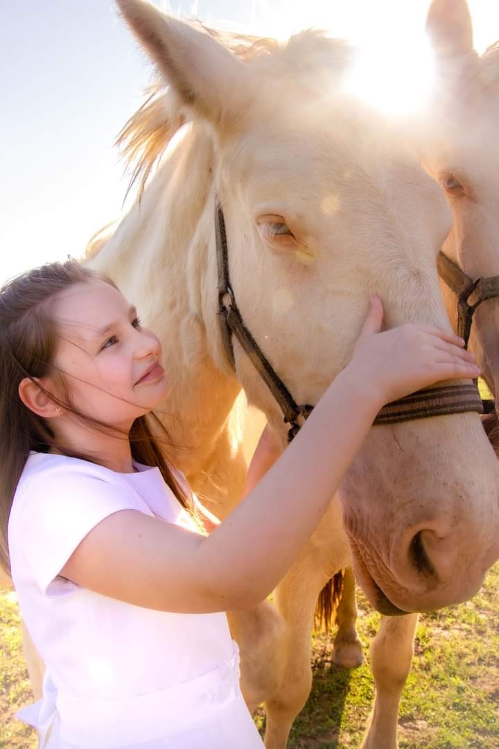 Dziewczynka w białej sukience czule dotyka pyska jasnego konia w uprzęży na tle słonecznej łąki.