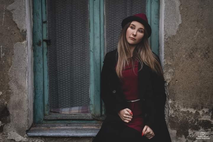 Portret kobiety w kapeluszu i płaszczu, opierającej się o starą, zieloną ramę okienną z szarą, teksturalną ścianą w tle.