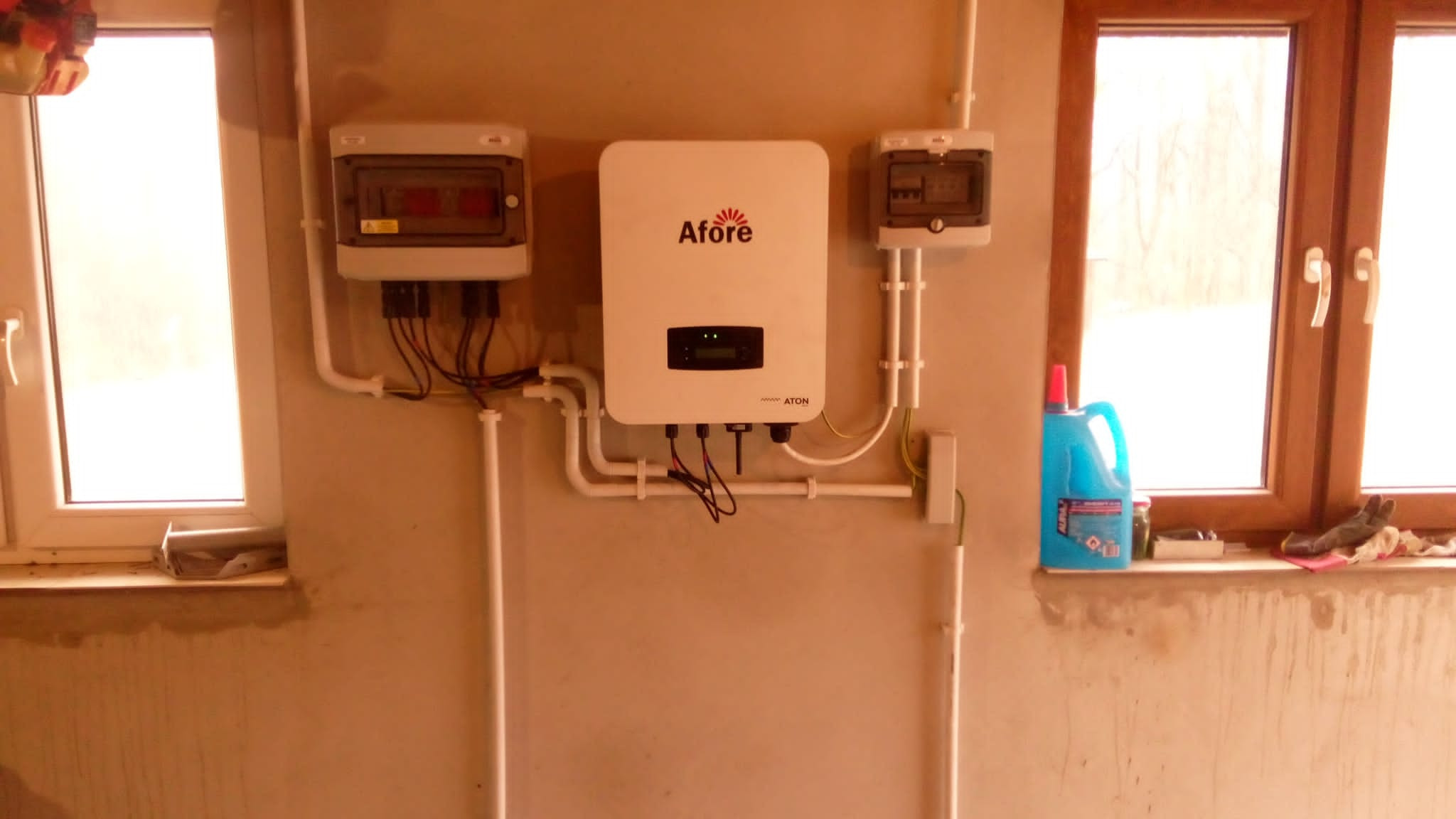 Centralnie umieszczony biały falownik firmy Afore z podłączonymi przewodami, obok dwa mniejsze panele elektryczne, wszystko zamontowane na ścianie w pomieszczeniu z dwoma oknami, na jednym...