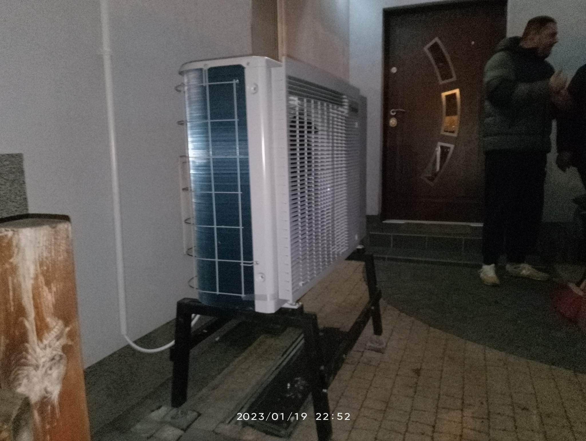 Klimatyzator typu split, jeszcze nie zamontowany, stoi na metalowym stojaku przed drzwiami wejściowymi do budynku.