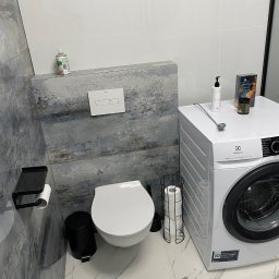 Akow - Nowoczesna łazienka z toaletą podwieszaną, białą pralką Electrolux i marmurową podłogą. Ściana za toaletą wykończona płytkami imitującymi abstrakcyjny obraz w odcieniach szarości i błękitu.