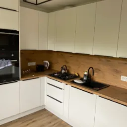 Nowoczesna kuchnia w bieli i drewnie: białe szafki z czarnymi uchwytami, drewniany blat i panel nad blatem, czarny zlew i bateria, płyta gazowa z czajnikiem, oświetlenie sufitowe w stylu...