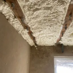 Ocieplenie poddasza pianką PUR, widoczne drewniane belki stropowe i metalowe uchwyty. Ściany surowe, okno zabezpieczone folią.