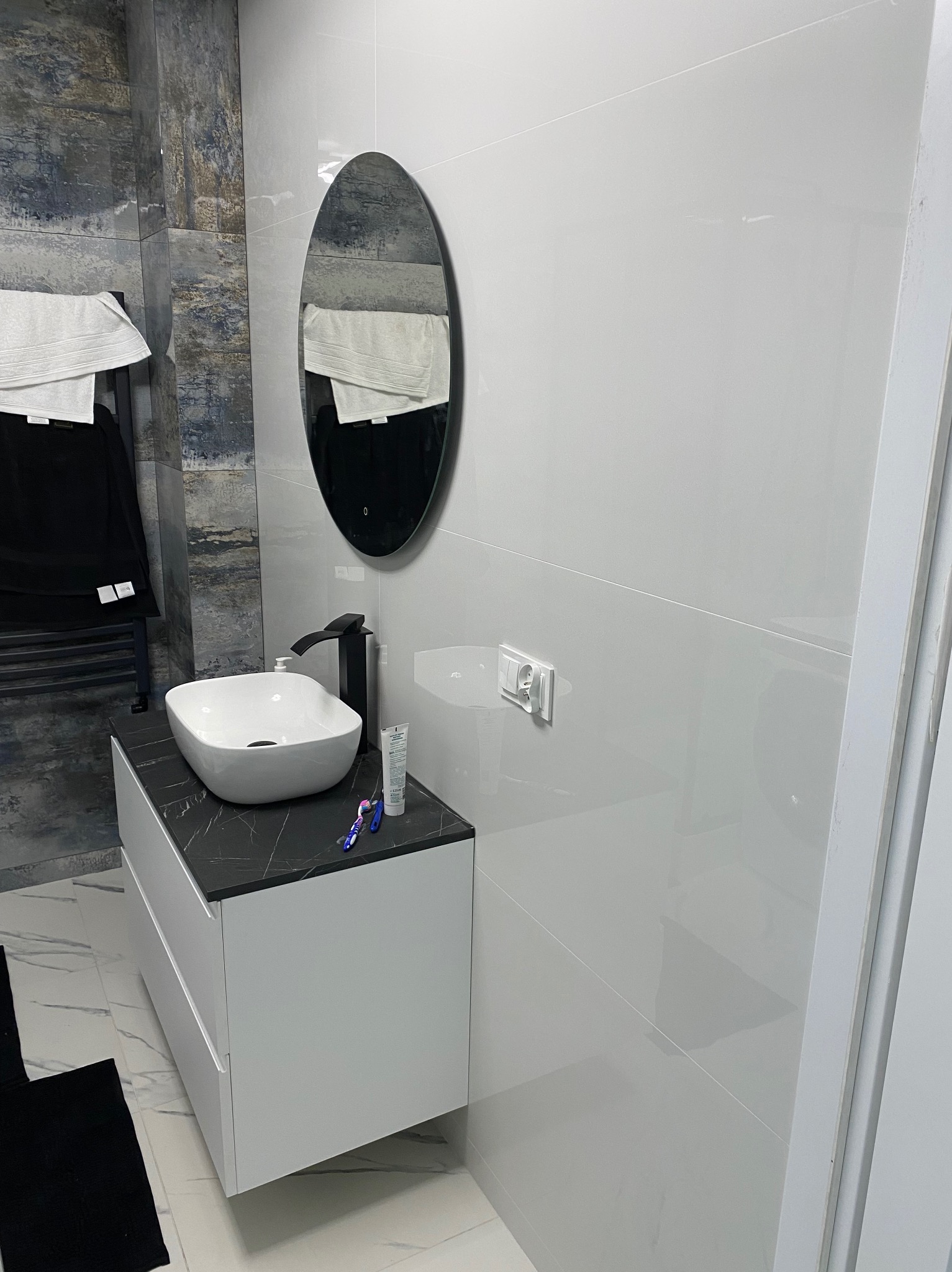 Nowoczesna łazienka z białą umywalką nablatową na szafce z czarnym blatem, owalne lustro w czarnej ramie, białe płytki ścienne i dekoracyjne płytki imitujące kamień, widoczny grzejnik drabinkowy...