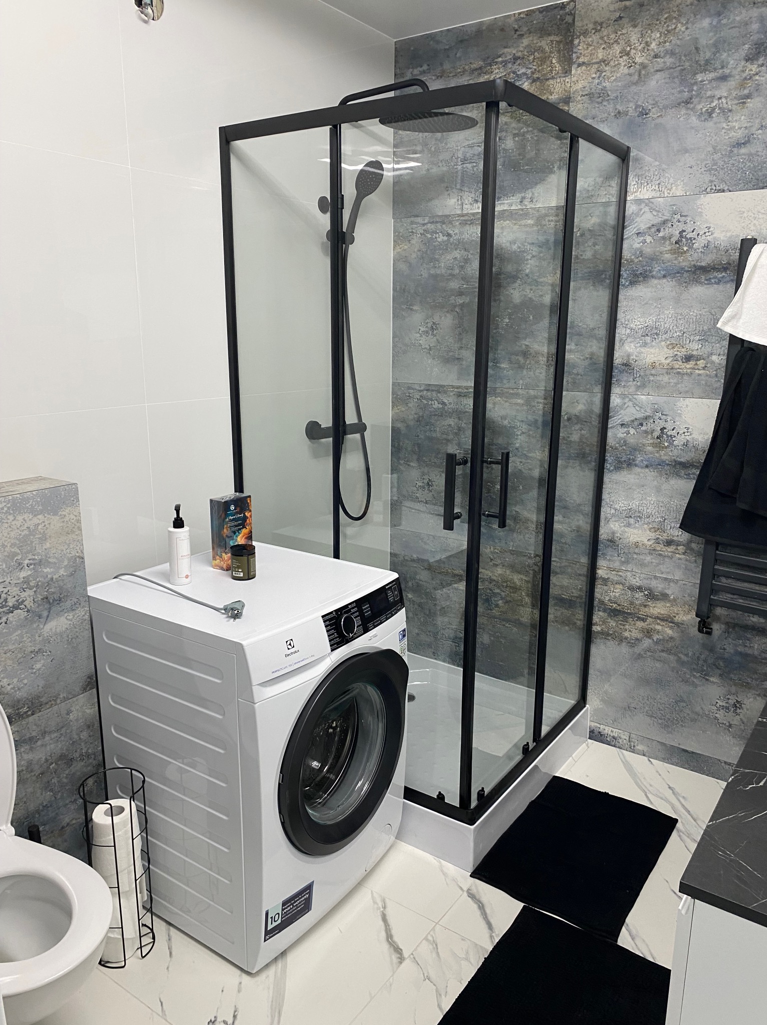 Nowoczesna łazienka z kabiną prysznicową typu walk-in z czarnymi profilami, pralką Electrolux i marmurowymi płytkami podłogowymi.