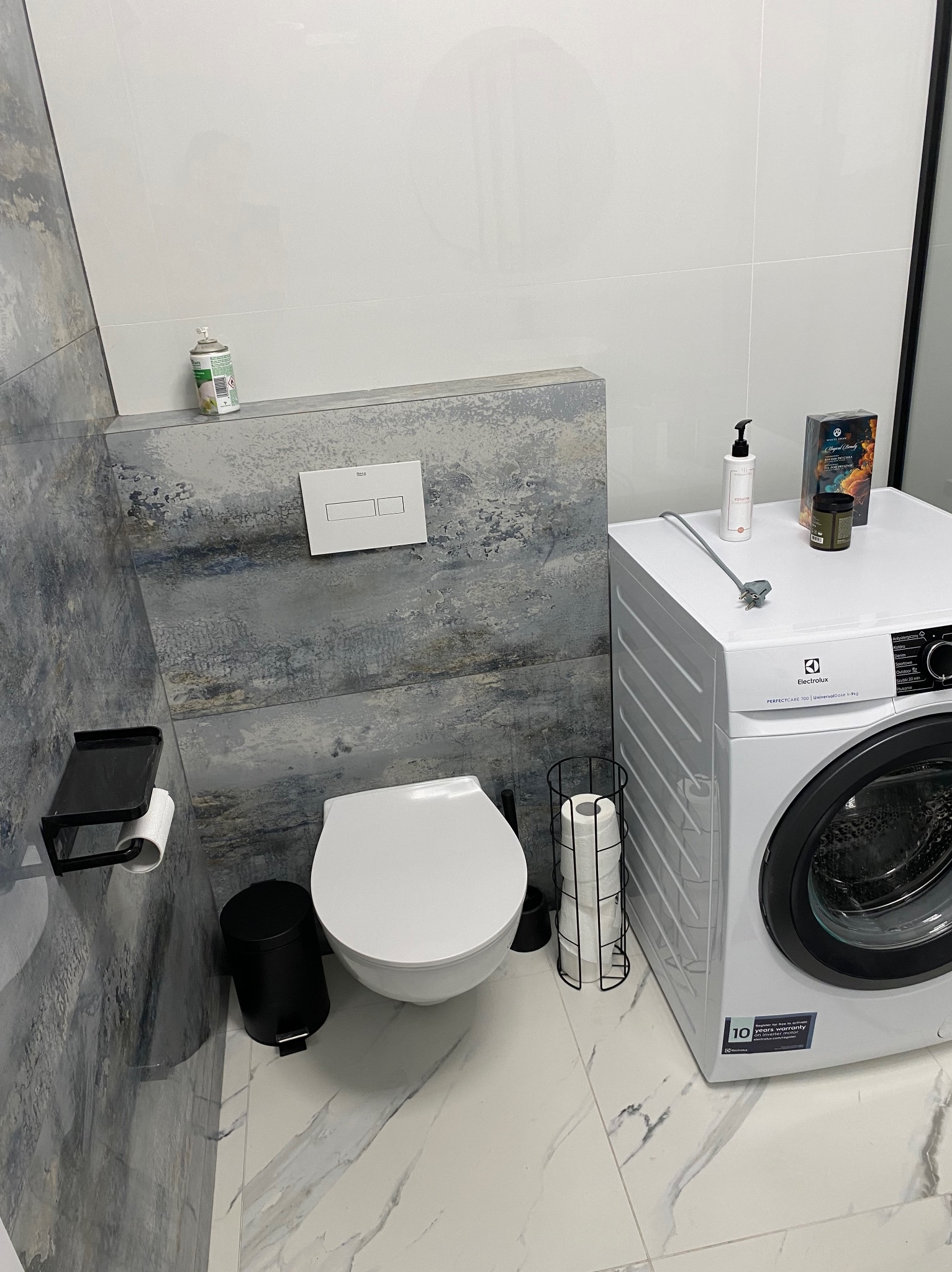 Nowoczesna łazienka z toaletą podwieszaną, białą pralką Electrolux i marmurową podłogą. Ściana za toaletą wykończona płytkami imitującymi abstrakcyjny obraz w odcieniach szarości i błękitu.