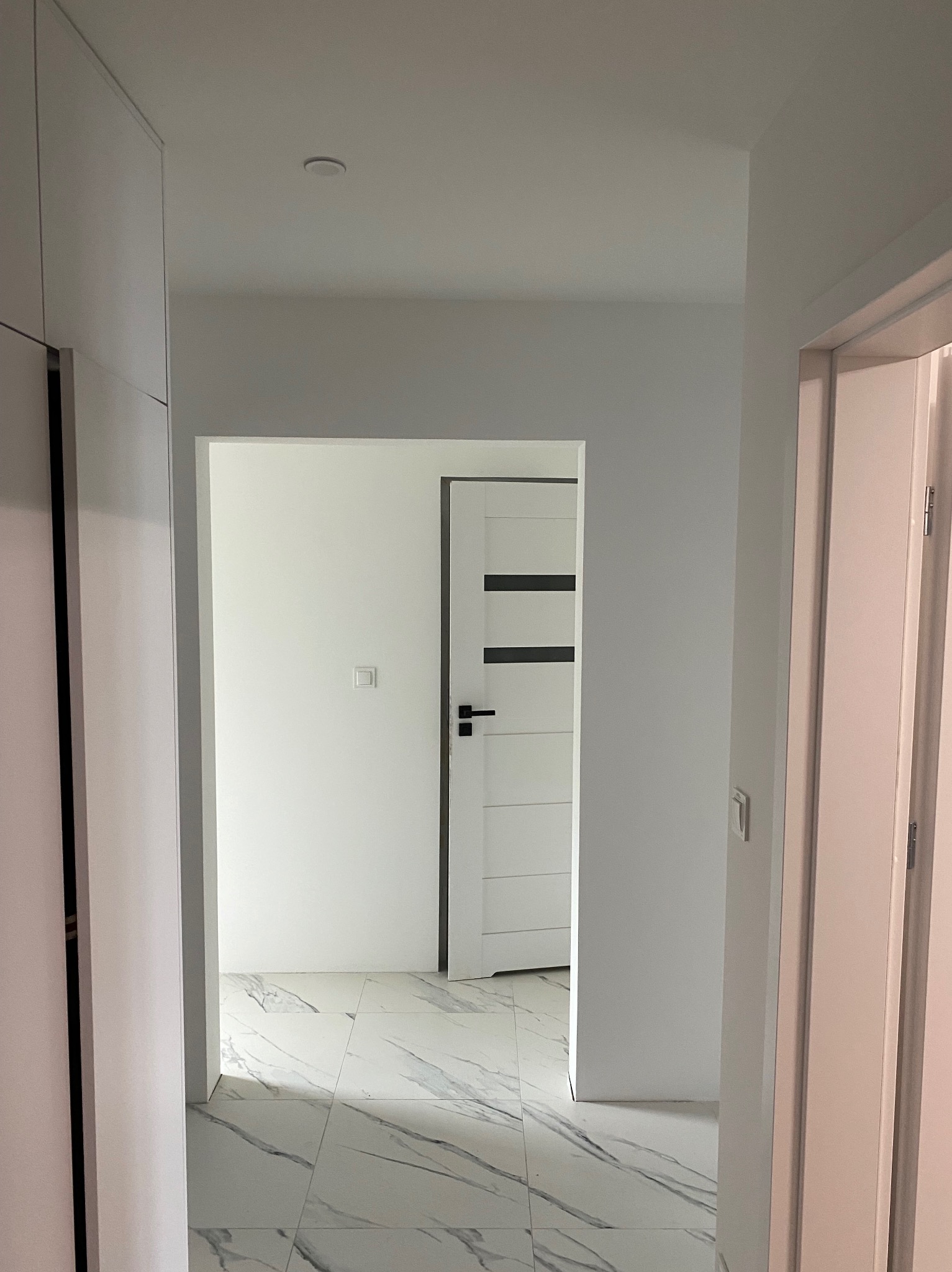 Nowoczesny korytarz z białymi ścianami, marmurową podłogą i minimalistycznymi drzwiami wewnętrznymi z czarnymi akcentami, widok z perspektywy wejścia.