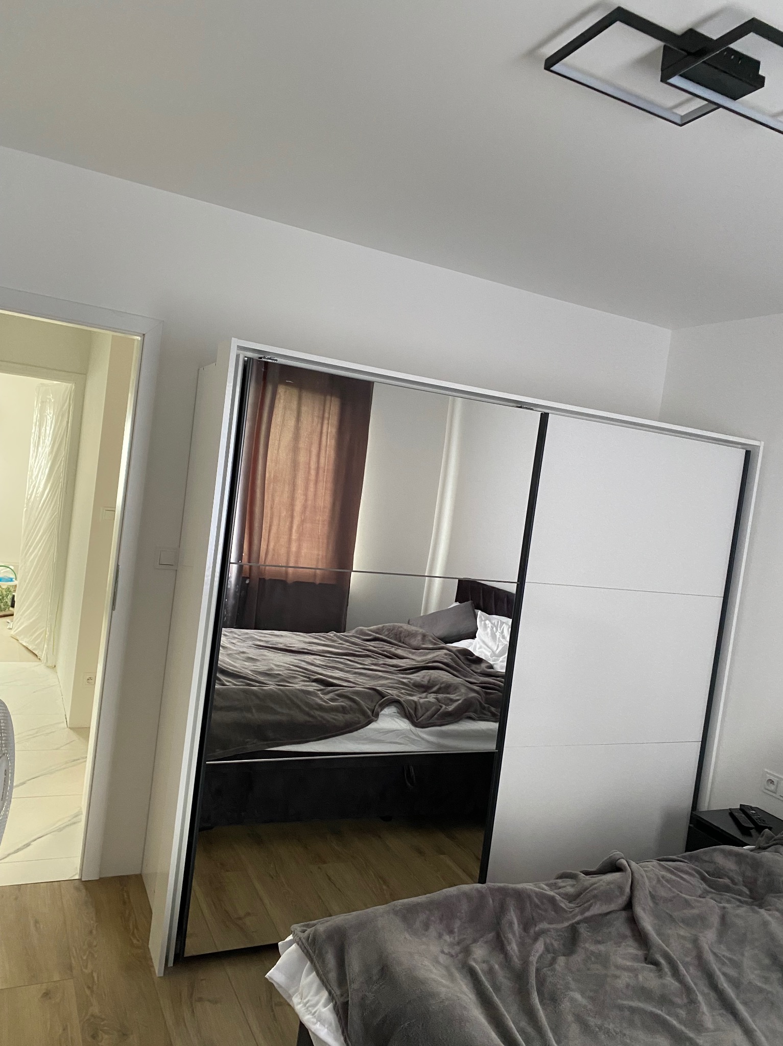 Nowoczesna szafa przesuwna z lustrzanymi drzwiami w sypialni, odbijająca łóżko i okno, z widocznym fragmentem sufitu z designerską lampą.