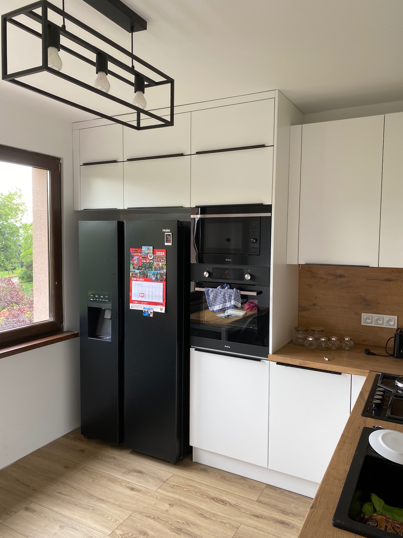 Nowoczesna kuchnia z białymi szafkami, czarną lodówką side-by-side z kalendarzem na drzwiach, piekarnikiem i mikrofalówką w zabudowie, drewnianym blatem i czarną lampą wiszącą